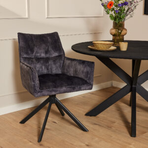 De Eetkamerstoel Dani | Velvet Antraciet, een moderne donkergrijze draaistoel met een zwart metalen onderstel, staat naast een zwarte ronde eettafel met kruispoot. De tafel staat vol met kleurrijke bloemen in een vaas, een gouden schaal en twee kopjes op een houten vloer.