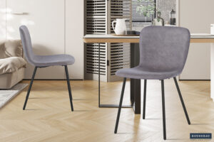Twee Stoel 'Susan' PU Taupe stoelen (set van 2) staan in een lichte, minimalistische eetkamer met een houten vloer, een tafel en een neutrale inrichting. Op de achtergrond zijn een bank en keukenkastjes te zien.