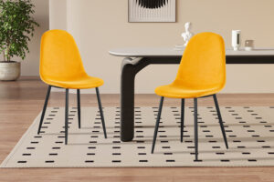Twee Stoel 'Boy' Fluweel Geel stoelen met zwarte poten staan naast een rechthoekige tafel in een minimalistische kamer met lichte muren, een tapijt met patroon, een potplant en decoratieve accenten op de achtergrond.
