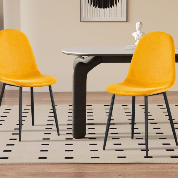 Twee Stoel 'Boy' Fluweel Geel stoelen met zwarte poten staan naast een rechthoekige tafel in een minimalistische kamer met lichte muren, een tapijt met patroon, een potplant en decoratieve accenten op de achtergrond.