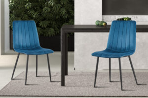 Twee moderne PROMO - Stoel 'Willis' Fluweel Blauw stoelen met zwarte poten staan rond een donkere, rechthoekige tafel op een licht tapijt, met groene bladplanten en een muur met marmereffect op de achtergrond.