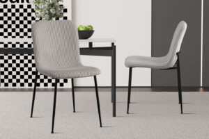 Twee Stoel 'Alice' Corduroy Lichtgrijs stoelen (set van 2) met zwarte metalen poten staan bij een witte tafel met een zwarte schaal met groene appels, in een lichte, minimalistische kamer met een licht tapijt en geblokte wandkunst.