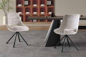 Twee Stoel 'Dante' Stof Beige stoelen (2 per doos) met zwarte metalen poten staan bij een strakke tafel op grijs tapijt. Op de achtergrond staan planken met witte kommen en boeken, plus een klein plantje ter decoratie.