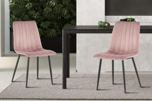 Twee roze fluwelen 'Willis' stoelen (Promo - Stoel 'Willis' Fluweel Roze) met zwarte poten staan naast een donkere houten tafel op een lichtgrijs kleed, met groene planten en een lichtgrijze muur op de achtergrond.
