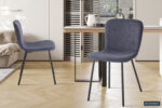 Twee Stoel 'Susan' Bouclé Donkergrijs stoelen (set van 2) met donkere metalen poten staan in een lichte eetkamer met een houten vloer, een houten tafel en een neutrale inrichting. Zonlicht vult de ruimte en creëert een gezellige sfeer.