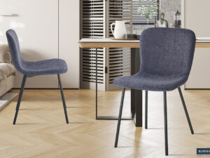 Twee Stoel 'Susan' Bouclé Donkergrijs stoelen (set van 2) met donkere metalen poten staan in een lichte eetkamer met een houten vloer, een houten tafel en een neutrale inrichting. Zonlicht vult de ruimte en creëert een gezellige sfeer.