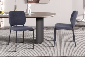 Twee moderne donkergrijze 'Sophia' stoffen stoelen staan op een lichtgestreept vloerkleed naast een ronde houten tafel. De achtergrond toont lichte kasten en minimalistische decoratie, waaronder een witte vaas en glas. (Set van 2, PROMO - Stoel 'Sophia' Stof Donkergrijs.).