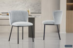Twee Stoel 'Arthur' Stof Ote Belfast 41 lichtgrijze stoelen (set van 2) met zwarte metalen poten staan op een glanzende tegelvloer in een moderne keuken met marmeren achterwand en neutrale kasten. Het Rousseau-logo staat rechtsonder.