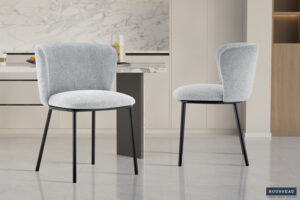Twee Stoel 'Arthur' Stof Ote Belfast 41 lichtgrijze stoelen (set van 2) met zwarte metalen poten staan op een glanzende tegelvloer in een moderne keuken met marmeren achterwand en neutrale kasten. Het Rousseau-logo staat rechtsonder.