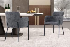 Twee Stoel 'Naomi' Stof Grijs fauteuils (2 per doos) staan op een licht tapijt aan een eettafel met wijn, glazen en brood. Minimalistisch decor en een winters buitentafereel zijn te zien door de grote ramen op de achtergrond.
