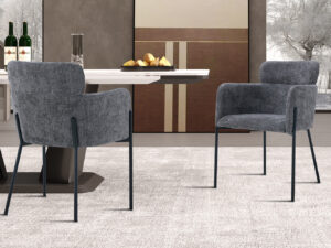 Twee Stoel 'Naomi' Stof Grijs fauteuils (2 per doos) staan op een licht tapijt aan een eettafel met wijn, glazen en brood. Minimalistisch decor en een winters buitentafereel zijn te zien door de grote ramen op de achtergrond.