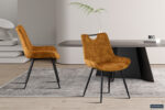 Twee elegante Eetkamerstoel 'Nina' Fluweel Goud stoelen (set van 2) met zachte goudkleurige fluwelen bekleding en zwarte metalen poten, geplaatst op een grijs vloerkleed in een moderne kamer met strak meubilair, een lichte houten vloer en een decoratieve plant.