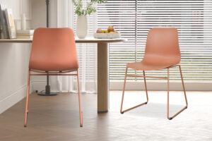 Twee Stoel 'Marie' PP Terracotta stoelen (2 per doos) met metalen poten staan bij een ronde houten tafel in een lichte kamer met grote ramen, witte jaloezieën en een vaas met witte bloemen op de plank.