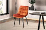 In een moderne eetkamer staat de Stoel 'Juta' Fluweel Oranje stoel met zwarte metalen poten op een vloerkleed met een licht patroon. Op de achtergrond staan een dressoir, vazen, een plant en een raam voor natuurlijk licht.