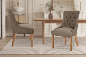 Twee Stoel 'Fancy' Stof Taupe stoelen (set van 2) met houten poten staan op een gestreept vloerkleed in een neutrale eetkamer, in combinatie met een houten tafel, open haard, vaas met takken en klassieke buste boven op een kast.