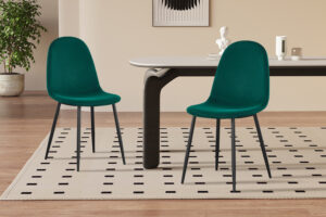 Twee Stoel 'Boy' Fluweel Groen stoelen (set van 2) met zwarte poten staan op een crèmekleurig vloerkleed met zwarte stippen, naast een moderne tafel in een minimalistische kamer met houten vloeren, een plant en abstract decor.