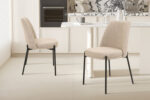 Twee 'Ella' stoelen in zandkleurige stof (SALE - Stoel 'Ella' Stof Zand, 2 per doos) met zwarte poten staan naast een witte keukentafel in een moderne keuken met marmeren achterwand en neutrale tinten.