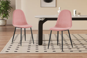 Twee Stoel 'Boy' Fluweel Roze stoelen met zwarte poten staan naast een rechthoekige tafel in een stijlvolle kamer met lichthouten vloeren, een vloerkleed met patroon, een plant en een minimalistische inrichting.