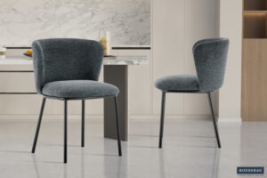 Twee moderne Stoel 'Arthur' Stof Ote Belfast 39 stoelen in donkergrijze stof (set van 2) met zwart metalen poten staan in een strakke keuken met marmeren achterwand, lichte kasten, gepolijste tegelvloer. Rousseau logo rechtsonder.