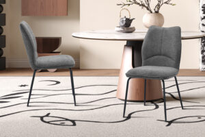 Twee Stoel 'Lara' Stof Ote Sea 456 Grijs stoelen (set van 2) met zwarte poten staan op een beige vloerkleed met abstracte zwarte lijnen, gepositioneerd bij een ronde tafel met houten onderstel in een eigentijdse eetkamer.