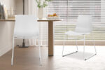 Twee Stoel 'Marie' PP Wit stoelen (2 per doos) met metalen poten staan in een lichte eethoek, samen met een ronde houten tafel met een fruitschaal en bloemen, naast grote ramen met horizontale jaloezieën.