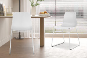 Twee Stoel 'Marie' PP Wit stoelen (2 per doos) met metalen poten staan in een lichte eethoek, samen met een ronde houten tafel met een fruitschaal en bloemen, naast grote ramen met horizontale jaloezieën.