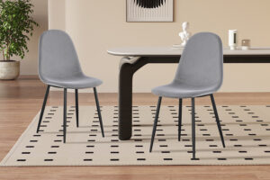 Twee Stoel 'Boy' Fluweel Grijs stoelen staan op een gestippeld tapijt naast een stijlvolle tafel in een moderne kamer met een houten vloer, een potplant en een minimalistische inrichting.