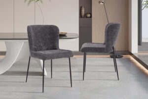 Twee Stoel 'Lisa' Bouclé Donkergrijs stoelen (2 per doos) met pluche donkergrijze stof en slanke zwarte metalen poten verfraaien een minimalistische eetkamer, in combinatie met een sculpturale witte tafel en decoratieve accenten op de achtergrond.