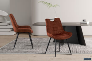 Twee Stoel 'Nina' Fluweel Roest stoelen met zwarte metalen poten staan op een grijs vloerkleed bij een strakke zwarte eettafel, geaccentueerd met minimalistische decoratie zoals een vaas met een plant en een kleine camera.