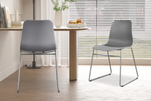 Twee Stoel 'Marie' PP Grijs stoelen (2 per doos) met modern design en metalen poten staan in een lichte kamer met houten vloer, een ronde fruittafel en grote ramen met luxaflex, die natuurlijk licht binnenlaten.