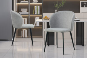 Twee Stoel 'Toledo' Stof Lichtgrijs stoelen (2 per doos) met zwarte metalen poten staan in een moderne keuken met glanzende vloeren, boekenplanken, een aanrecht met een schaal sinaasappels en een stijlvolle inrichting.