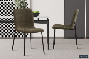 Twee moderne, groene Stoel 'Alice' Bouclé stoelen (set van 2) met zwarte metalen poten staan rond een strakke witte tafel met een zwart onderstel. Een schaal met groene appels staat op tafel, met abstracte muurkunst op de achtergrond.