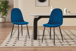 Twee Stoel 'Boy' Fluweel Donkerblauw stoelen met zwarte poten staan naast een rechthoekige tafel op een beige vloerkleed met zwarte blokjes, in een stijlvolle kamer met houten vloeren, een plant en decoratieve accessoires op de achtergrond.