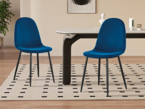 Twee moderne donkergrijze fluwelen 'Juta' stoelen (2 per doos) staan op een licht vloerkleed met een zwart vierkant patroon, naast een rechthoekige tafel in een minimalistische kamer met houten vloeren, een plant en decoratieve items op de achtergrond.