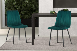 Twee Stoel 'Willis' Fluweel Groen stoelen met zwarte poten staan op een lichtgrijs vloerkleed naast een donkerhouten tafel, omringd door groene planten en decoratieve objecten op de achtergrond.