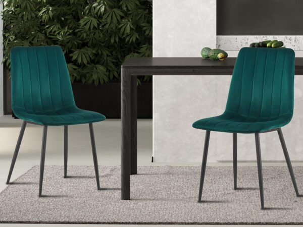 Twee Stoel 'Willis' Fluweel Groen stoelen met zwarte poten staan op een lichtgrijs vloerkleed naast een donkerhouten tafel, omringd door groene planten en decoratieve objecten op de achtergrond.