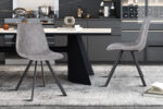 Twee Stoel 'Emma' PU Donkergrijs stoelen (2 per doos) met metalen poten staan op een gedessineerd vloerkleed in een stijlvolle keuken met donkere kasten en een tafel met boeken en decor op de achtergrond.