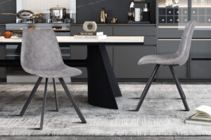 Twee Stoel 'Emma' PU Donkergrijs stoelen (2 per doos) met metalen poten staan op een gedessineerd vloerkleed in een stijlvolle keuken met donkere kasten en een tafel met boeken en decor op de achtergrond.