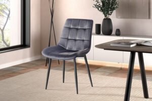 Een moderne kamer met een Stoel 'Juta' Fluweel Taupe fluwelen stoel (set van 2) met metalen poten op een beige gedessineerd tapijt, bij een raam, naast een tafel met een boek, decoratieve vazen en een grote plant op de achtergrond.