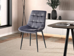 Een moderne kamer met een Stoel 'Juta' Fluweel Taupe fluwelen stoel (set van 2) met metalen poten op een beige gedessineerd tapijt, bij een raam, naast een tafel met een boek, decoratieve vazen en een grote plant op de achtergrond.