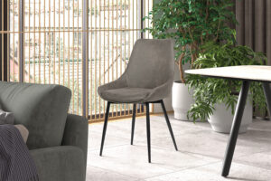 Een moderne woonkamer met de Stoel 'Mirano' Micro fiber PU Grijs stoel (2 per doos) op zwarte poten, geplaatst bij een grijze bank, ronde tafel, weelderige groene potplanten en hoge ramen met houten verticale lamellen voor natuurlijk licht.