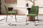 Twee Stoel 'Lara' Stof Ote Sea 360 groene stoelen staan op een licht vloerkleed met abstracte zwarte lijnen bij een ronde marmeren eettafel met een houten onderstel. Op de tafel staat een decoratieve vaas.