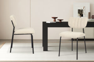 Twee Stoel 'Antura' Ote Lisa 110 Beige stoelen (2 per doos) met zwarte poten staan op een licht vloerkleed voor een zwarte tafel, terwijl minimalistische kandelaars en een ingelijste print het dressoir op de achtergrond sieren.