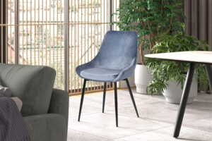 Een moderne woonkamer met Stoel 'Mirano' Fluweel Blauw accentstoelen op zwarte poten, geplaatst bij een ronde tafel en groene planten in potten. Hoge ramen met verticale jaloezieën vullen de ruimte met natuurlijk licht.