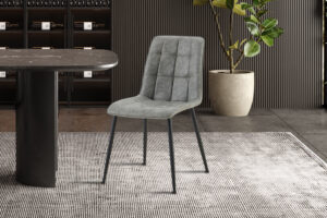 Een set van 4 'Furas' Vintage PU Grijs stoelen heeft een moderne grijze bekleding en slanke zwarte poten, gerangschikt naast een ronde zwarte tafel op een vloerkleed met textuur, met een grote potplant en een wand met houten panelen op de achtergrond.