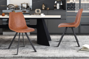 Twee moderne Stoel 'Emma' PU Cognac stoelen met zwarte metalen poten staan naast een strakke zwarte eettafel in een eigentijdse keuken met grijze kasten, een marmeren achterwand en een licht vloerkleed met patroon. Boeken en decor staan op tafel.