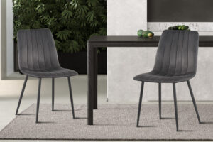 Twee moderne 'Willis' grijs fluwelen stoelen staan aan een donkere houten tafel op een licht vloerkleed, met een groene bladplant en decor op de achtergrond.