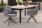 Twee Stoel 'Lisold' Draaibaar tot 0 Stof Fabb Raven 13 Donkergrijs stoelen (set van 2) staan bij een lichtstenen rechthoekige eettafel in een moderne eetkeuken met grote ramen en natuurlijk licht.
