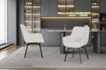 Twee Stoel 'Modest' Draaibaar Stof Fabb Raven 22 Ecru fauteuils staan op een groot vloerkleed voor een strakke, donkergrijze keuken met ingebouwde planken, flessen, glaswerk en onderkastverlichting. Op tafel staat een plant in een glazen vaas.