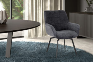 De PROMO - Stoel 'Modest' Draaibaar Stof Fabb Raven 18 Donkergrijs voegt moderne stijl toe met zijn donkergrijze bekleding en metalen poten, geplaatst op een blauw vloerkleed naast een ronde tafel in een lichte kamer met uitzicht op tropische planten.